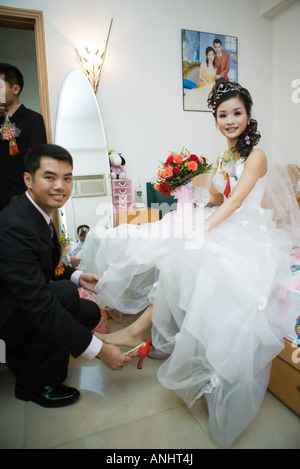 Groom putting shoe on bride's foot in bedroom, both smiling at camera, full length Stock Photo