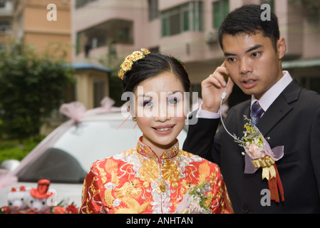 Bride wearing traditional Chinese clothing, smiling at camera, groom behind using earphone Stock Photo