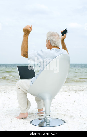 Senior man using laptop and cell phone on beach, raising arms Stock Photo