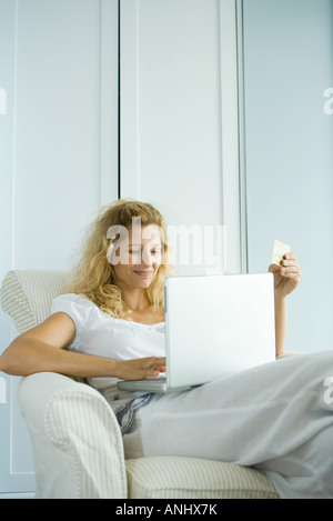 Woman making online purchase with credit card, sitting in armchair with laptop on lap Stock Photo