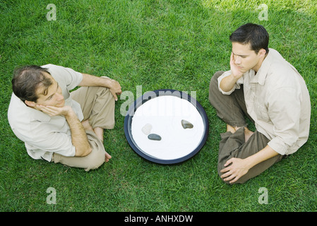 Two men sitting on the ground next to rock garden, both with hands under chins, high angle view Stock Photo
