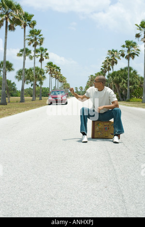 Man sitting on suitcase in the middle of street, looking over shoulder at approaching car Stock Photo