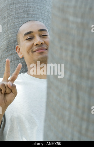 Man leaning against tree trunk, making peace sign, smiling at camera Stock Photo