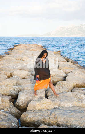 A colour portrait image of a beautiful dark haired woman walking along a rocky outcrop in the resort of Puerto Pollensa,Majorca. Stock Photo