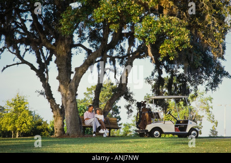 Senior couple sitting on golf bench with golf cart nearby Stock Photo