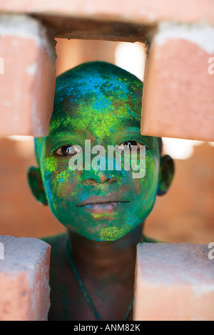 Young Indian boy covered in coloured powder pigment looking through a cross hole in a  brick building. India Stock Photo