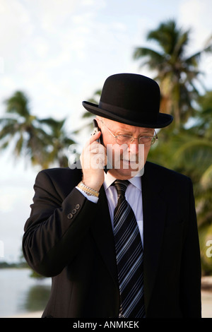 Maldives workaholic businessman in bowler hat and business suit talking on mobile phone walking along tropical beach Stock Photo