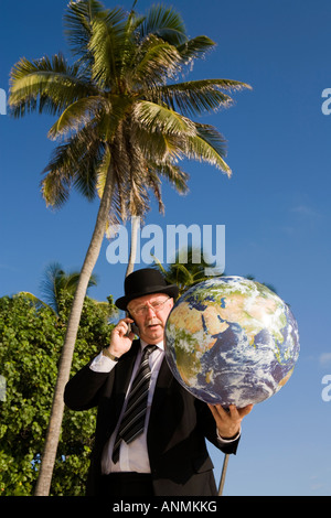 businessman on beach in bowler hat and business suit with globe in palm of hand talking on mobile phone Stock Photo