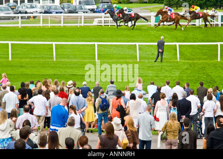 Horizontal view of a crowd of spectators standing against the rails watching horses and jockeys racing passed on a sunny day Stock Photo