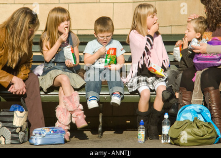 Horizontal close up of two mums with young children having a picnic lunch on a park bench in the sunshine Stock Photo