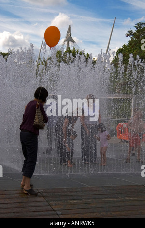 a mother who is holding an orange ballon looks on a her children play in a modern fountain with some of her friends Stock Photo