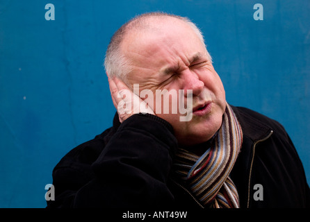 middle aged balding man wearing a scarf suffering from ear ache - wincing in pain as he cups his ear with his hand - Stock Photo