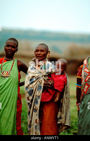 A Maasai women dressed in traditional clothing stands with her young ...