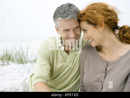 Mature couple sitting on beach, smiling with heads together Stock Photo