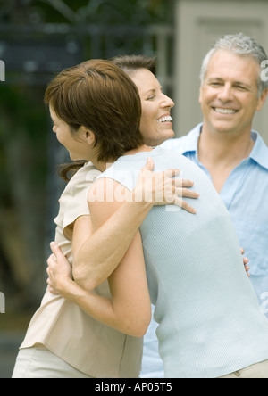 Two mature women hugging while man smiles in background Stock Photo