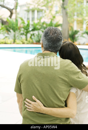 Couple standing with arms around each other, looking at swimming pool Stock Photo
