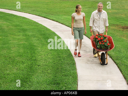 Father and adult daughter walking on walkway pushing wheelbarrow full of flowers Stock Photo