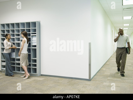 Office scene, two women talking by mailboxes while man using cell phone walks through hallway