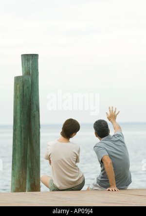 Couple sitting on edge of dock, man waving Stock Photo