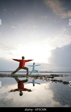 Man and woman doing yoga exercises The Warrior, reflection in the water, Buchscharn, lake Starnberg, Bayern, Germany Stock Photo