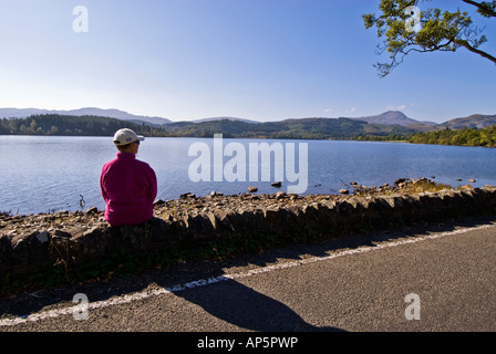 Retired tourist sitting on a stone wall gazing over the loch to Ben Lomond in the distance Stock Photo