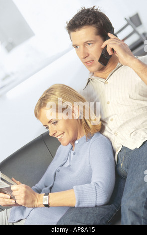 Young man using a mobile phone with a young woman reading a newspaper on a sofa Stock Photo