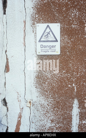 Rendered ochre wall with large cracks and weathered white paint and bleached yellow sign with icon and Danger Fragile roof Stock Photo