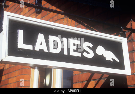 Old-fashioned carved wooden sign in black and white hanging from orange brick wall with pointing hand stating Ladies