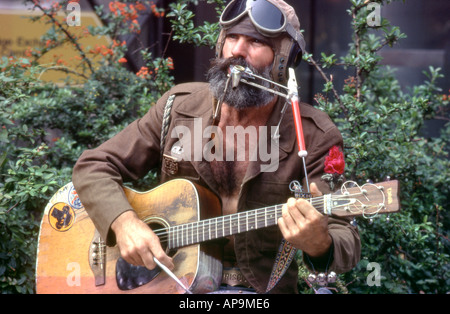 A hairy bearded 1970s American busker man musician playing guitar wearing aviator goggles performing in the street  Cologne Germany 1977  KATHY DEWITT Stock Photo