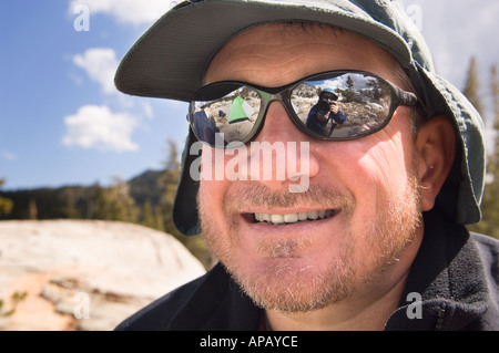 face of a smiling middle aged man with a goatee in a hiking cap and sunglasses reflecting a tent and a photographer Stock Photo