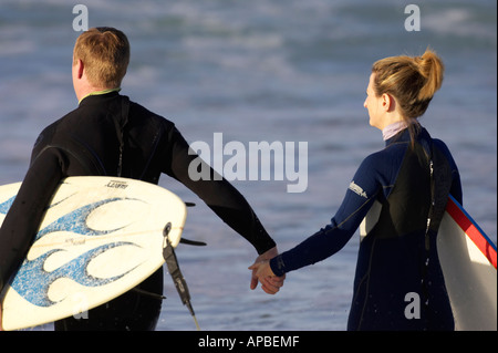 male and female surfers in wetsuits hold hands walking out to sea off white rocks beach portrush county antrim Stock Photo