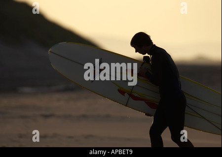 silhouette of man in wet suit carrying surf board out of the sea at the end of the day Stock Photo