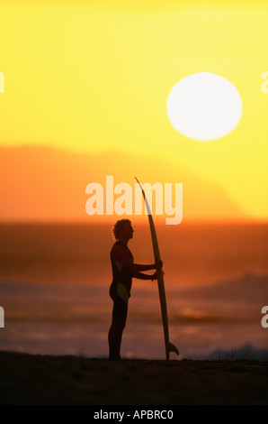 Surfer standing with board on beach at sunset Stock Photo