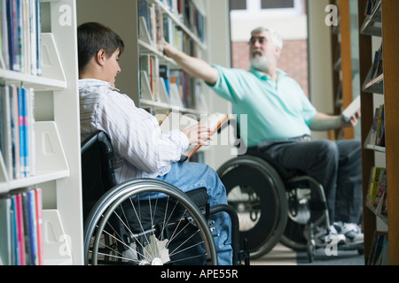 Middle-aged man disabled man picking a book from a bookshelf with a mid adult woman reading a book in a library Stock Photo
