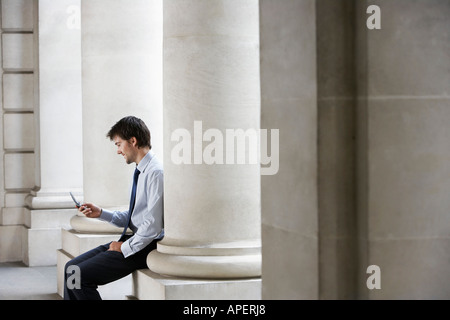 Businessman outside building sitting on pillar sending text message Stock Photo