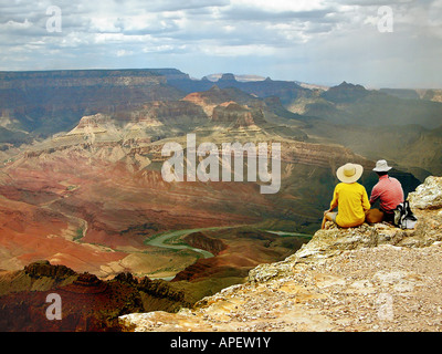 Couple sitting on edge of precipice overlooking the Grand Canyon with Colorado River in the distance. Stock Photo