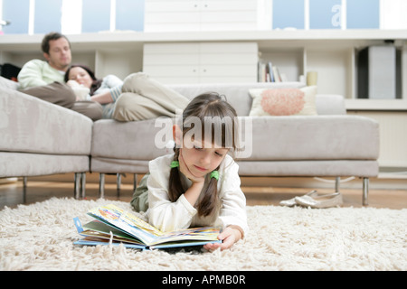 Girl reading book on floor, parents on sofa in background Stock Photo