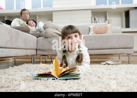 Girl reading book on floor, parents on sofa in background Stock Photo