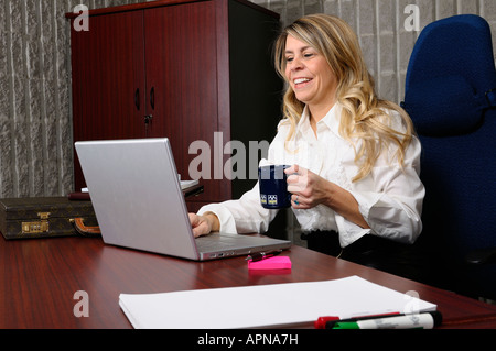 Smiling female executive in office with coffee mug working on a laptop computer at desk Stock Photo