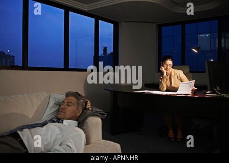 Businesswoman Working Late, Man Sleeping on Sofa in Office Stock Photo