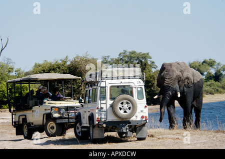Tourists in two game viewing vehicles watching a bull elephant emerge from a waterhole. Stock Photo