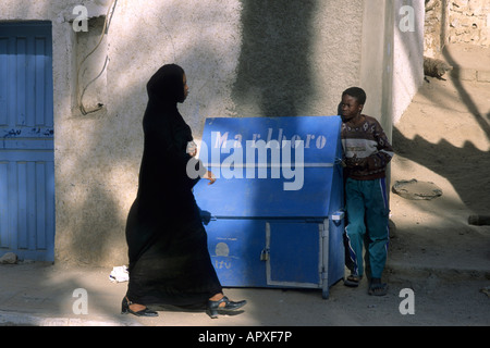 Woman wearing traditional garment walks past young cigarette seller on the streets of Djanet Stock Photo