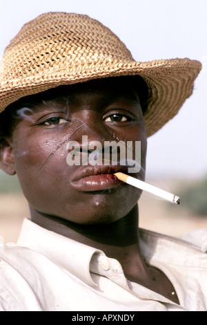 Portrait of a young Burkina Faso man with tribal scarring on his face wearing a straw hat and smoking a cigarette Stock Photo