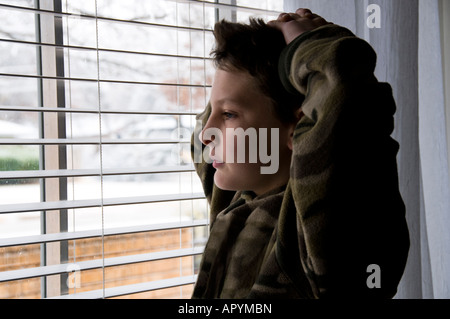 A 13-year-old Caucasian boy appearing sad & lonely stands in front of a window looking out. USA. Stock Photo