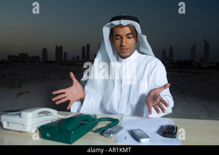 Busy Young Arab Businessman sitting at a desk with Dubai City in the background with lots of telephones Stock Photo