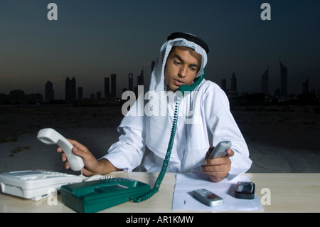 Busy Young Arab Businessman sitting at a desk with Dubai City in the background with lots of telephones Stock Photo