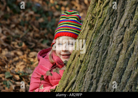 small child with red jacket and hat, hides behind a tree trunk Stock Photo