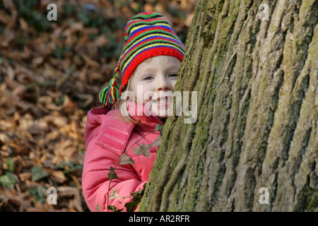 small child with red jacket and hat, hides behind a tree trunk Stock Photo