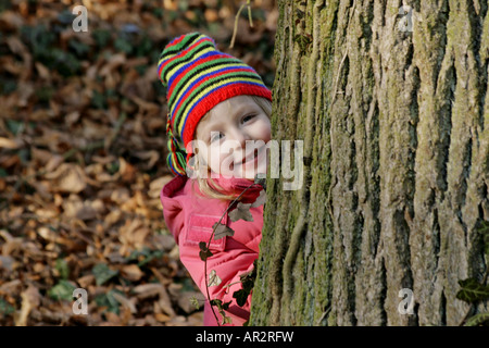 small child with red jacket and hat, hides behind a tree trunk Stock Photo