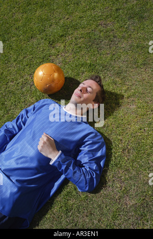 Young man wearing football shirt lying on grass next to a golden coloured football Stock Photo